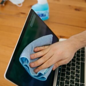 A person wipes a laptop screen using a blue cloth and spray bottle on a wooden table for hygiene.